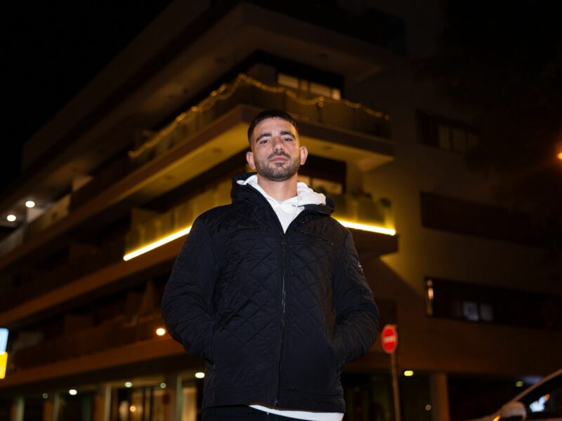 אדם עומד עם מעיל שחור בלילה עם וברקע בניין מגורים מואר