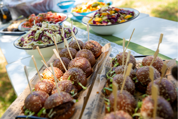 "מגש עם מיני־בורגרים על שיפודים ושתי קערות סלט על שולחן פיקניק בחוץ."