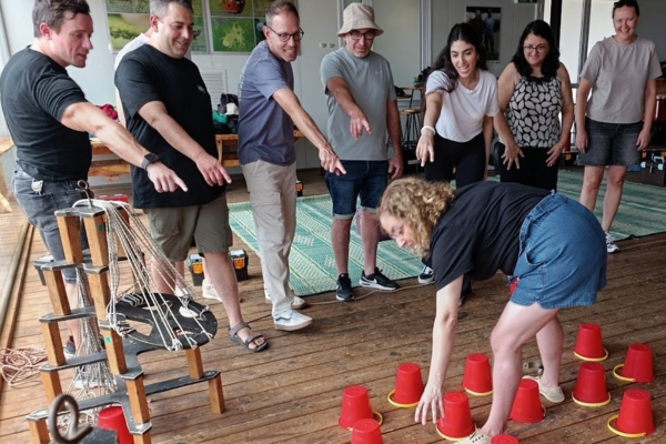 "קבוצה של משתתפים מצביעים ומכוונים משתתפת שמנווטת בין כוסות משחק אדומות על רצפת עץ; משמאל מתקן חבלים."