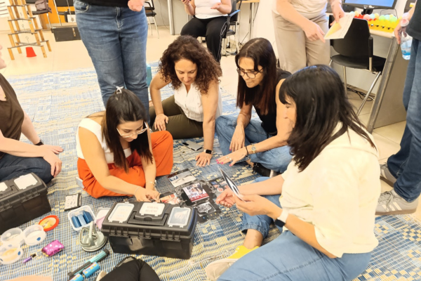 "קבוצה של משתתפות יושבות על שטיח בכיתה וממיינות חלקים וערכות כלים מתוך קופסאות."