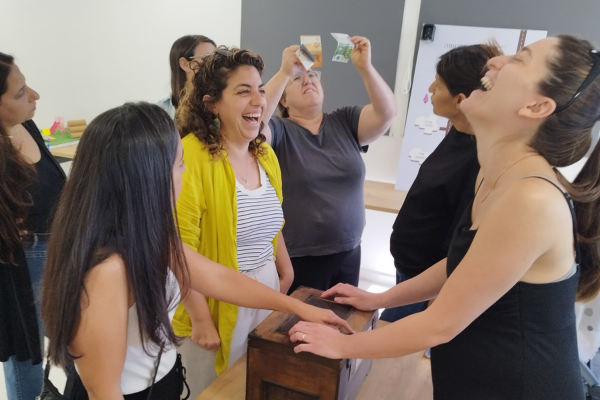 "קבוצה של משתתפות צוחקות סביב קופסת עץ בכיתה; שתיים מניחות ידיים על הקופסה, וברקע משתתפת מחזיקה שטרות."
