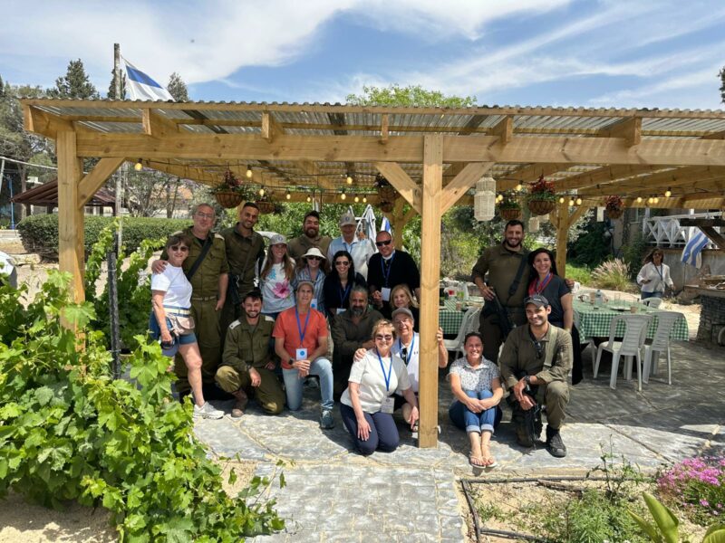 "חיילים ומתנדבים מצטלמים יחד תחת פרגולה מעץ בגינת אירועים; שולחנות ערוכים ודגלי ישראל ברקע."