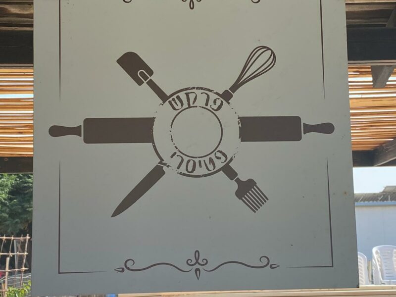 שלט תלוי עם איור מערוך במרכז וכלי מטבח מצטלבים סביבו—מרית, מטרפה, סכין ומברשת—מעל שולחנות עץ בחוץ."